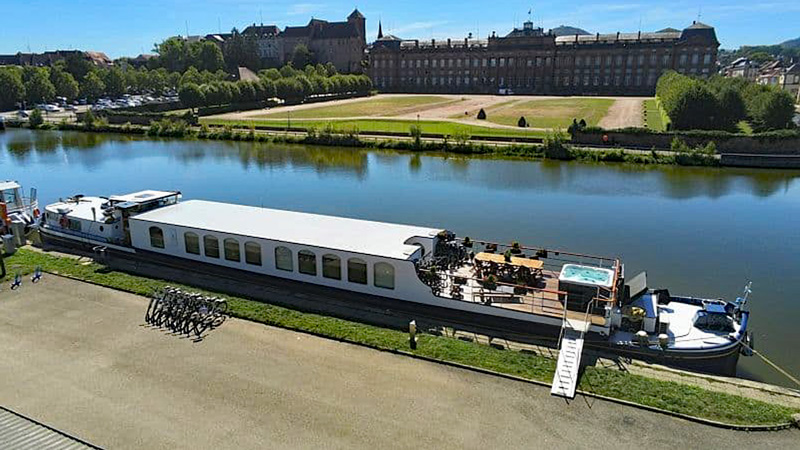 Luxury Hotel Barge Panache moored along the canal in Alsace France. Drone view of entire barge with gangplank, sundeck with hot tub, table & chairs, with bicycles on land next to the boat