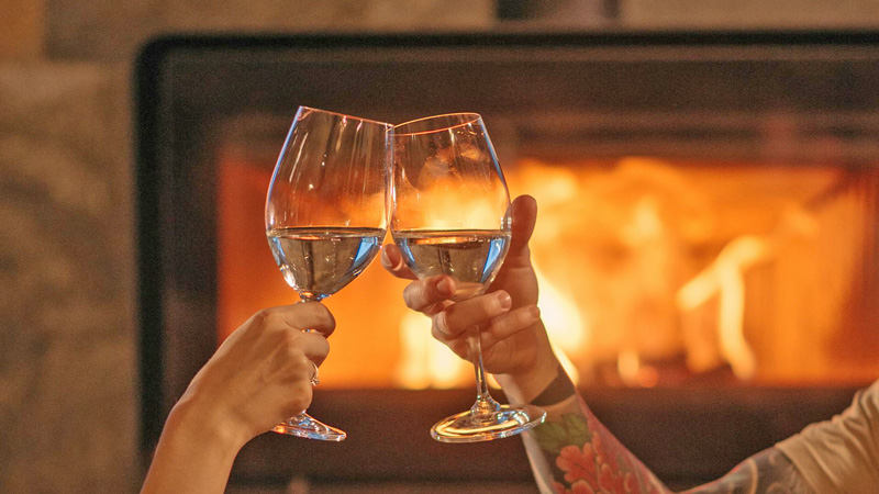 Cheers! Toast of two glasses of white wine in front of a fireplace. Photo: Pexel - DanilyUK